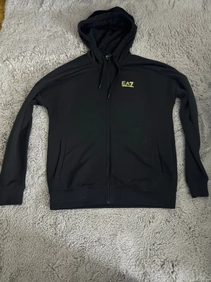 Svart EA7 hoodie med dragkedja - Säljer en svart hoodie från EA7 med gul logga på bröstet. Knappt använd. En ny går för ungefär 1200kr