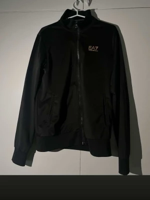 Svart zipjacka från EA7 Emporio Armani - Snygg svart zipjacka från EA7 Emporio Armani med guldfärgad logga på bröstet och dekorativa ränder i nacken. Jackan har hög krage, två fickor med dragkedja och ribbade muddar. Perfekt för en sportig och stilren look.