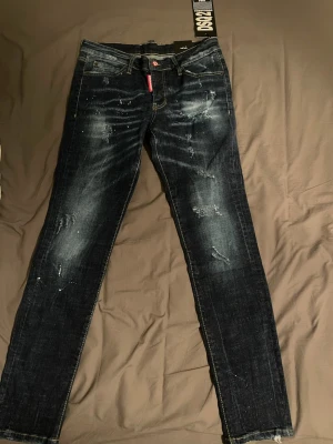 Mörkblå Dsquared2 jeans med slitningar - Säljer ett par mörkblå jeans från Dsquared2 med coola slitningar och distressed detaljer. Jeansen har raka ben, klassisk femficksmodell och en röd logotagg vid gylfen. Materialet är denim i bomull och passformen är slim.