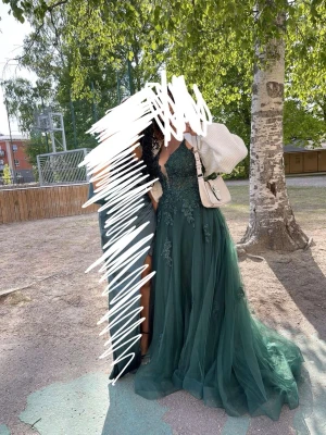 Mörkgrön långklänning med spetsdetaljer - Elegant mörkgrön långklänning med v-ringning och spetsdetaljer på överdelen och öppen rygg. Klänningen har tunna axelband och ett flytande, voluminöst kjolparti i flera lager tyg. Perfekt för bal eller festliga tillfällen.