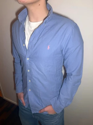 Blå skjorta från Ralph Lauren - Klassisk blå skjorta från Ralph Lauren med button-down krage och rosa broderad logga på bröstet. Skjortan har långa ärmar, vita knappar och är gjord i mjuk bomull. Perfekt för en clean och stilren look.