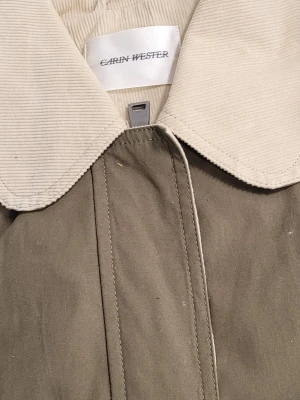 Grön jacka med kontrastkrage från Carin Wester Khaki Green - Stilren grön jacka Funktionsjacka AMELA  I Khaki Green från Carin Wester med stor, ljus krage i manchester och två stora fickor framtill. Jackan har en rak passform och dold knäppning för en clean look. Perfekt för dig som gillar minimalistisk stil.