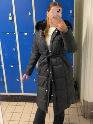 Svart lång dunjacka med huva och bälte - Snygg svart lång dunjacka med tjock huva och knytbälte i midjan. Jackan har elastiska muddar, varm vaddering och knäppning framtill. Perfekt för kalla vinterdagar och ger en stilren look. Fickorna har tyvär små hål i sig den ena är större en den andra, fickorna funkar oavsett men kan vara lite jobbigt, jag kan ha tur om jag hittar nån som gillar o sy, osen är snöret tyvär borta men jackan funkar fortfarande super bra priset kan även diskuteras 