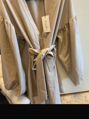 Beige kappa med knytskärp - Trendig beige kappa från Bik Bok med vida ärmar och knytskärp i midjan. Kappan har en avslappnad passform och djup V-ringning. Perfekt för dig som gillar stilrena och moderna plagg. Materialet känns mjukt och följsamt.