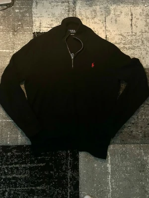 Svart halvzip tröja från Polo Ralph Lauren - Klassisk svart långärmad tröja från Polo Ralph Lauren med halv dragkedja och röd broderad logga på bröstet. Tröjan har hög krage och är tillverkad i ett mjukt stickat material. Perfekt för en stilren och avslappnad look.