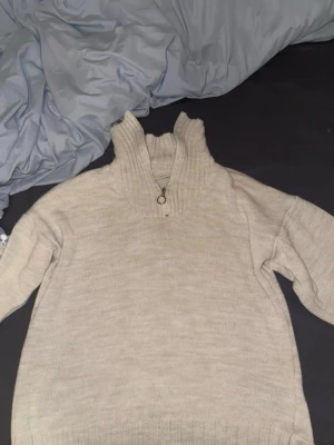 Beige stickad tröja med halv dragkedja - Mysig beige stickad tröja med hög krage och halvzip-dragkedja framtill. Tröjan har långa ärmar och ribbade detaljer vid halsen. Nyskick 