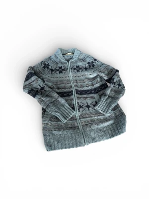 Kofta - Mysig gråblå stickad kofta med dragkedja och nordiskt stjärnmönster i mörkblått och grått. Koftan har ribbade muddar, hög krage och är perfekt för kalla dagar. Materialet ser ut att vara ullblandning och modellen är rak och avslappnad.