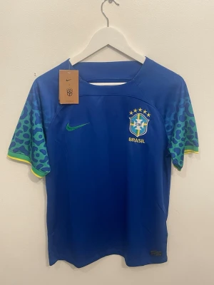 Nike Brasil  - Säljer en officiell Brasilien fotbollströja från Nike i blått med gröna och blå leopardmönstrade ärmar och gula detaljer. Tröjan har CBF-logga, Nike Swoosh och Dri-FIT-teknologi för extra komfort. Perfekt för dig som älskar fotboll och vill sticka ut på planen. PRIS KAN DISK