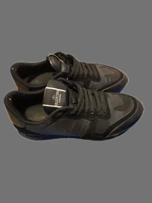 Svarta sneakers från Valentino - Snygga svarta sneakers från Valentino med kamouflagemönster och svarta skosnören. Skorna har en låg siluett och är tillverkade i en mix av läder och textil. Perfekta för dig som vill ha en stilren och trendig look.