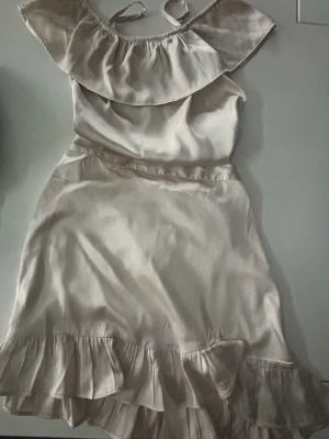 Champagnefärgad offshoulder volangklänning - Snygg kortklänning i champagnefärgad satin med offshoulder och volangdetaljer både upptill och nertill. Klänningen har smala axelband och en lös, flowy passform som ger en romantisk vibe. Perfekt för sommarens varma dagar.tröjan har storlek 146/152 och kjolen har storleken 158/164