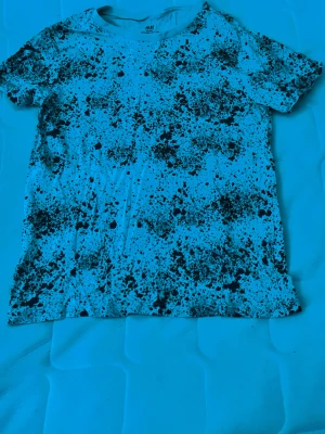 Vit t-shirt med svart splatter från H&M - Säljer en vit t-shirt från H&M med svart splattermönster. Tillverkad i ekologisk bomull och har en klassisk rund hals och korta ärmar. Perfekt för dig som gillar en unik och kreativ stil.