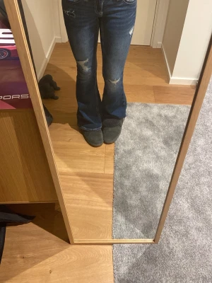 Blå bootcut jeans med broderi - Säljer ett par blå bootcut jeans med slitningar och snyggt vitt broderi på bakfickorna. Jeansen har låg midja och klassisk femficksmodell. Perfekta för dig som gillar en avslappnad men trendig stil.