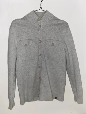 Ljusgrå kofta med bröstfickor - Stilren ljusgrå kofta med knäppning framtill och två bröstfickor med lock och knapp. Hög ribbad krage och ribbade muddar vid ärmslut och nederkant. Perfekt för lager på lager och enkel att matcha med olika outfits.