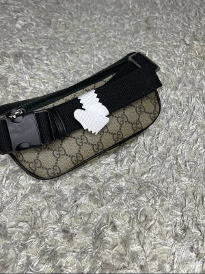 Gucci axelväska med monogram - Snygg axelväska från Gucci med klassiskt GG-monogram i beige och brunt tyg. Väskan har svarta läderdetaljer och en bred svart justerbar axelrem med spänne. Perfekt för dig som gillar streetstyle och designeraccessoarer.