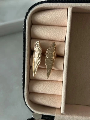 Guldfärgad ring med änglavingar - Unik guldfärgad ring med två detaljerade änglavingar som motiv. Ringen har en öppen design och är tillverkad i metall med blank finish. Perfekt för dig som gillar smycken med symbolisk touch och vill sticka ut lite extra.