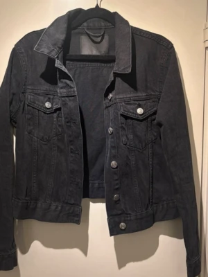 Svart jeansjacka med silverknappar - Stilren svart jeansjacka med klassisk krage och silverfärgade knappar framtill. Två bröstfickor med lock och knapp, samt markerade sömmar för en snygg look. Perfekt att slänga över en hoodie eller t-shirt för en avslappnad vibe. Helt ny, pris kan diskuteras 