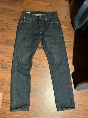Mörkblå Levi's jeans 501 - Mörkblå jeans från Levi's i klassisk femficksmodell med raka ben. Jeansen har kontrastsömmar, guldfärgade nitar och Levi's ikoniska läderpatch bak i midjan. Tillverkade i kraftig denim för en stilren och tidlös look. Helt spritt nya. Säljer då de är för små.