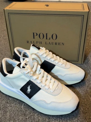 Vita sneakers från Polo Ralph Lauren - Stilrena vita sneakers från Polo Ralph Lauren med svart detalj och logga på sidan. Skorna har en klassisk låg siluett, vita snören och en vit sula med svart yttersula. Materialet är en mix av mocka och textil för en modern look.