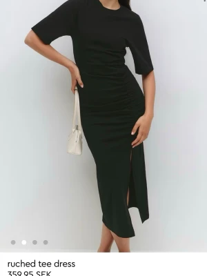 Gina tricot ruched tee dress - Stilren svart klänning i mjukt material med korta ärmar, rynkade detaljer längs sidan och en snygg slits framtill. Klänningen har en rak passform och enkel rund halsringning, perfekt för en trendig och avslappnad look. Storlek M, prislappen är kvar. Slutsåld på hemsidan 