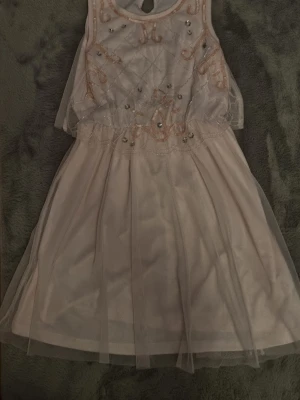 Ljusrosa barn klänning med paljettdetaljer - Superfin ljusrosa klänning från Kappahl med glittriga paljettdetaljer och broderier på överdelen. Klänningen har ärmlös design, rund halsringning och ett lager av tunn tyll som ger en drömmig look. Perfekt för speciella tillfällen! Använd ett par gånger och har lite skavanker på paljettdetaljerna. Skadorna syns inte mycket. Säljer pga för liten.