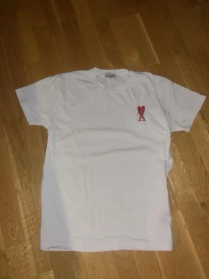 Vit t-shirt från Ami Paris med hjärtdetalj - Säljer en vit t-shirt från Ami Paris med en röd broderad hjärtdetalj och bokstaven A på bröstet. Klassisk passform och rund halsringning. Perfekt basplagg med en snygg detalj som sticker ut.