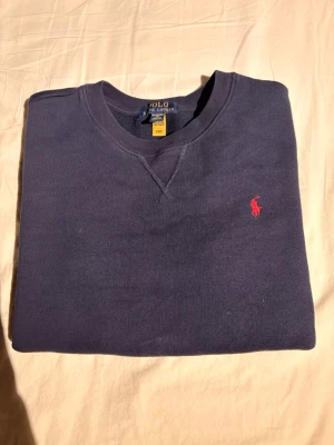 Mörkblå sweatshirt från Polo Ralph Lauren - Mörkblå sweatshirt från Polo Ralph Lauren med klassisk rund halsringning och röd broderad logga på bröstet. Tröjan har långa ärmar och ribbade muddar vid ärmslut och nederkant. Perfekt för en avslappnad och stilren look.