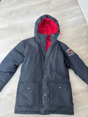 Svart Helly Hansen dunjacka med röd insida - Snygg svart dunjacka från Helly Hansen med röd vadderad insida och huva. Jackan har två stora fickor framtill med knappar och dragkedja, samt Helly Hansen-logga på ärmen. Perfekt för kalla vinterdagar.