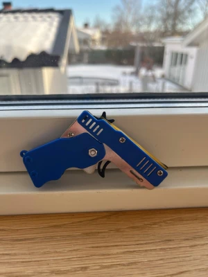 Blå miniatyr gummibandspistol - Säljer en liten gummibandspistol i metall och plast. Pistolen är blå med detaljer i silver och svart samt några gula inslag. Perfekt för dig som gillar små mekaniska prylar eller vill ha något kul att skjuta gummiband med.