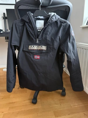 Svart vindjacka från Napapijri - Snygg svart vindjacka från Napapijri med halv dragkedja, huva och stor ficka framtill. Jackan har Napapijri-logga och norsk flagga på bröstet samt patch på ärmen. Perfekt för blåsiga dagar och streetwear-stil.