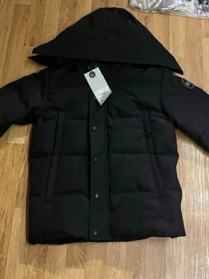 Svart pufferjacka från Canada Goose - Snygg svart pufferjacka från Canada Goose med stor huva och klassisk logotyp på ärmen. Jackan har breda kanaler, tryckknappar framtill och två stora fickor. Perfekt för kalla dagar och riktigt varm. Materialet är slitstarkt och vattenavvisande.