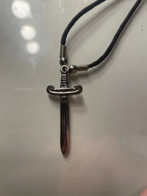 Svart halsband med svärdhänge - Coolt halsband med svart snöre och ett silverfärgat svärdhänge. Hänget har detaljerad design som liknar ett klassiskt svärd och snöret har metallfärgade detaljer vid fästena. Perfekt accessoar för dig som gillar edgy stil.
