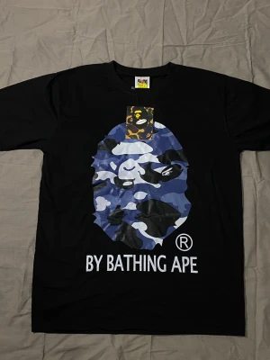 BAPE TSHIRT - Säljer en svart t-shirt från By Bathing Ape med det ikoniska apansiktet i blått, vitt och svart camomönster på bröstet. Stor logga under trycket. Klassisk passform och rund hals. Perfekt för dig som gillar streetwear och vill sticka ut.