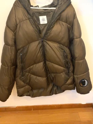 Olivgrön pufferjacka från C.P. Company - Säljer en olivgrön pufferjacka från C.P. Company med ikoniska glasdetaljer på huva och ärm. Jackan har quiltad design, dragkedja framtill och två fickor med dragkedja. Perfekt för kalla dagar och ger en riktigt streetwear-vibe.