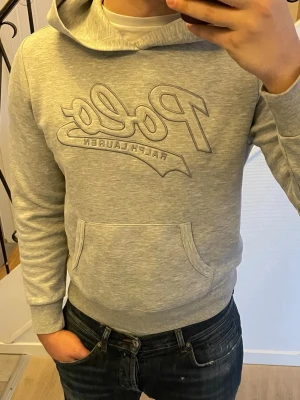 Grå hoodie från Polo Ralph Lauren - Snygg grå hoodie från Polo Ralph Lauren med stor broderad logga på bröstet. Klassisk känguruficka framtill och ribbade muddar vid ärmar och midja. Tillverkad i mjukt bomullsmaterial och har en skön huva. Perfekt för en avslappnad och trendig look.