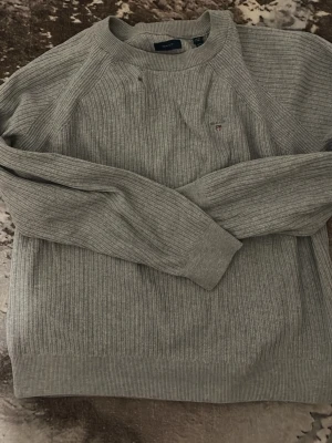 Grå ribbstickad tröja från Gant - Snygg grå ribbstickad tröja från Gant med rund halsringning och diskret logga broderad på bröstet. Tröjan har långa ärmar och breda muddar vid ärmslut och nederkant. Perfekt för lager på lager och enkel att matcha med olika outfits.