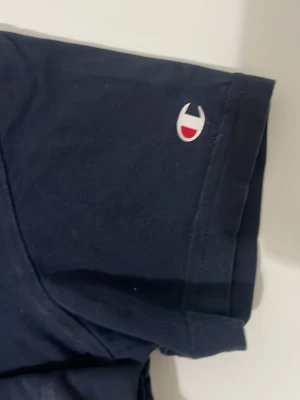 Mörkblå Champion t-shirt - Snygg mörkblå t-shirt från Champion med klassisk logga på ärmen. T-shirten har korta ärmar och är gjord i mjuk bomull, perfekt för en avslappnad stil.