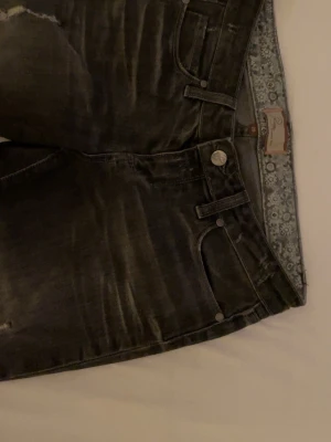 Svarta slitna jeans  - Säljer ett par svarta jeans med slitningar och trasiga detaljer på benen. Jeansen har klassisk femficksmodell, knappgylf och en snygg tvättad look. Perfekta för dig som gillar en lite tuffare stil.