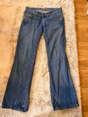 Blå bootcut jeans från Replay - Säljer ett par klassiska blå bootcut jeans från Replay med fem fickor och låg midja. Jeansen har en snygg tvätt och NY äpple logga i rött på bakfickan. Perfekta för dig som gillar retrostil och vill ha ett par jeans med lite vidare ben. Köpte dem från tinlessel. Mått: midjemått:40cm midjehöjd:18 cm innebenslängd:79cm ytterbenslängd:99cm benöppning:25cm 