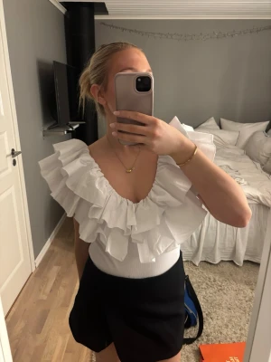 Vit volangbody med djup v-ringning - Storlek L men passar mer M! Supersnygg vit body med stora volanger och djup v-ringning. Toppen är ärmlös och har en figurnära passform som framhäver axlarna. Perfekt för dig som vill sticka ut med en trendig och feminin look. Har ett litet hål i ryggen, men går att sy igen. 
