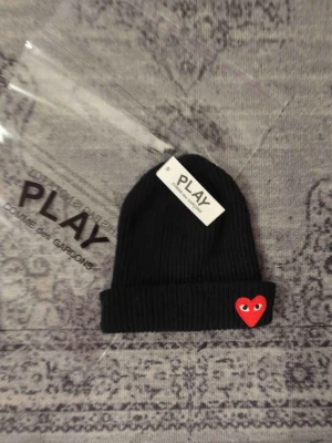 Svart mössa Comme des Garçons Play - Svart ribbstickad mössa från Comme des Garçons Play med uppvikt kant och den ikoniska röda hjärtloggan med ögon framtill. Perfekt accessoar för dig som gillar streetwear och vill ha en stilren men ändå lekfull detalj. Materialet känns mjukt och stretchigt.