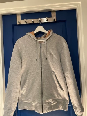 Grå zip hoodie från Burberry - Snygg grå zip hoodie från Burberry med klassiskt rutigt foder i huvan. Hoodien har dragkedja framtill, två stora fickor och justerbar dragsko i huvan. Perfekt för dig som gillar stilrena plagg med en twist av lyx. Materialet är mjukt och bekvämt, perfekt för vardagslooken.