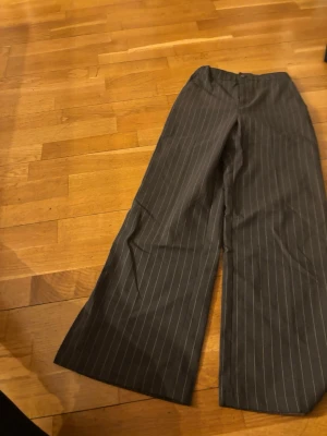 Gråa kostymbyxor med kritstreck - Säljer ett par gråa kostymbyxor med kritstrecksrandigt mönster och vida ben. Byxorna har hög midja och klassisk stängning fram. Perfekta för en stilren och trendig look.