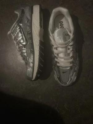 Nike silverfärgade sneakers - Säljer ett par riktigt snygga Nike sneakers i silver och vitt. Skorna har en futuristisk design med reflekterande detaljer, vita snören och en chunky sula. Perfekta för dig som vill sticka ut och gillar sportig streetstyle. Modellen har tydliga Nike-loggor på sidan och tungan.