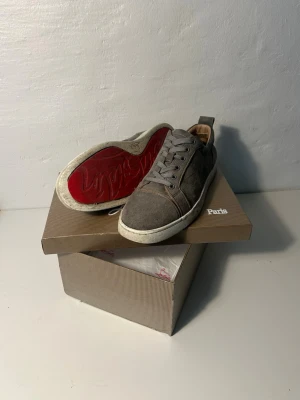 Gråa Christian Louboutin sneakers - Snygga gråa sneakers från Christian Louboutin med klassisk låg siluett och signaturröd sula. Skorna är gjorda i mocka med gråa skosnören och vita kanter runt sulan. Perfekta för dig som vill ha en stilren och exklusiv look. 