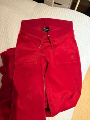 Röda velourbyxor Juicy Couture - Säljer ett par klassiska röda mjukisbyxor från Juicy Couture i mjuk velour. Byxorna har dragsko i midjan, stora fickor fram och en bakficka med knappar. Ikonisk JC-logga broderad på ena fickan. Perfekta för en chill och trendig look.