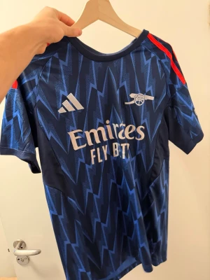 Blå Arsenal fotbollströja Adidas - Snygg blå Arsenal fotbollströja från Adidas med blixtmönster, röda detaljer på axlarna och tryckta loggor framtill. På ryggen står det EZE och nummer 10 i vitt. Tillverkad i lätt och ventilerande material, perfekt för träning eller match.