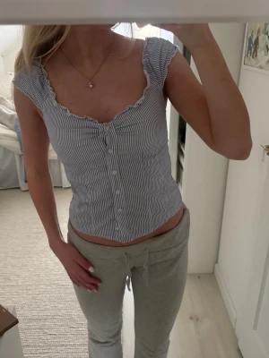 Randig blus med volangkant - Söt blus med smala blå och vita ränder, knappar framtill och volangkant vid ärm och hals. Modellen är croppad och har en feminin, figurnära passform. Perfekt för dig som gillar en romantisk och trendig stil.