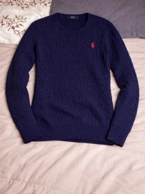 Mörkblå kabelstickad tröja Polo Ralph Lauren - Kabelstickad mörkblå tröja från Polo Ralph Lauren med klassisk röd logga broderad på bröstet. Tröjan har rund halsringning, lång ärm och ribbade muddar. Perfekt till höst och vinter för en stilren look.  PRIS KAN DISKUTERAS!