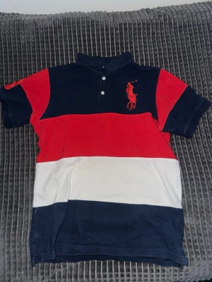 Randig pikétröja från Polo Ralph Lauren - Säljer en snygg pikétröja från Polo Ralph Lauren med breda ränder i marinblått, rött och vitt. Tröjan har klassisk krage, knappar framtill och den ikoniska polospelaren broderad på bröstet. Perfekt för en avslappnad men stilren look.
