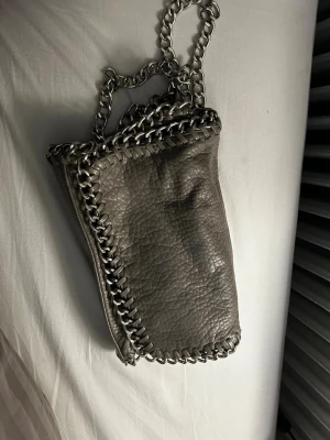 Grå kedjeväska i veganskt läder - Snygg grå handväska med grov kedja och detaljerade sömmar längs kanterna. Väskan har magnetlås och innerfack med dragkedja. Tillverkad i veganskt läder med en cool, lite skrynklig textur. Perfekt för dig som gillar edgy accessoarer.
